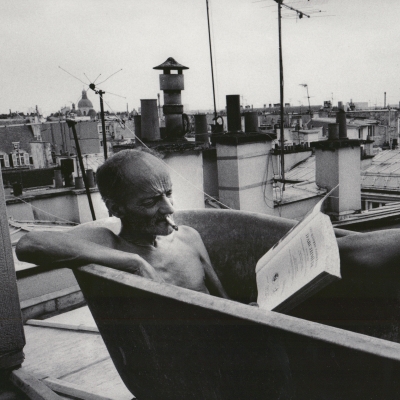 A man sits in a rooftop bathtub smoking and reading a book